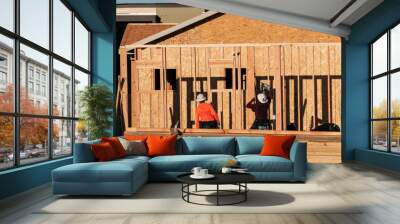 In a housing development in Queen Creek, Arizona, two wood framers wearing hard hats are using power equipment, such as a pneumatic nailer, to construct the wooden framework of a tract home Wall mural