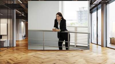 Portrait of a confident businesswoman in business suit holding a cup of coffee while using cellphone during break-time in the business building. Business stock photo. Wall mural