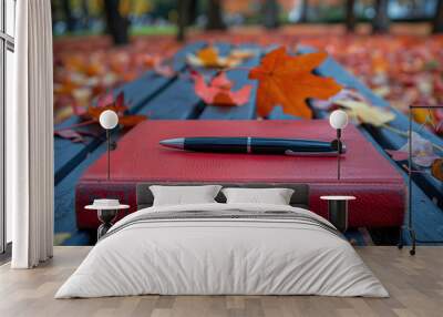 Pen lying on red notebook on a bench covered with autumn leaves Wall mural