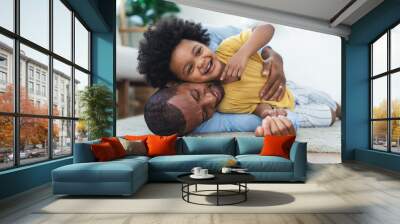 Black father in living room have a fun playing together on warm floor at home.Happy African family dad hugging, embracing his lovely child while lying on the floor.Sweet moments of fatherhood concept. Wall mural