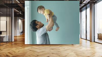 Asian Father lifting baby in yellow shirt and grey leggings in the air against blue wall, both smiling, Candid family moment, love and bonding concept. Wall mural