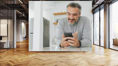 Attractive Middle-aged man using mobile phone or smartphone while standing in kitchen at home. Wall mural