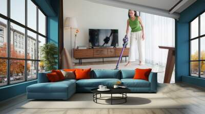 Young Asian Woman vacuuming a living room rug with a cordless vacuum cleaner for housekeeping housework and chores in a modern home. Wall mural