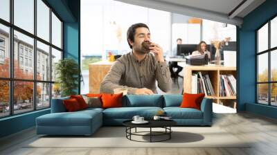 Hispanic man with mustache eating and enjoying a chocolate sweet bun. Unhealthy habits in the office concept. Wall mural