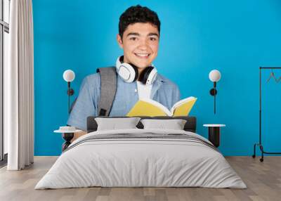 Portrait of hispanic college student holding a book isolated on blue background. Wall mural