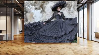 A young woman in a black gown and a wide-brimmed hat poses against a weathered wall. Wall mural