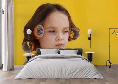 a 5-year-old girl with curlers and hair clips in her hair on a yellow isolated background. A little girl was twisting orange curlers around her hair. Wall mural