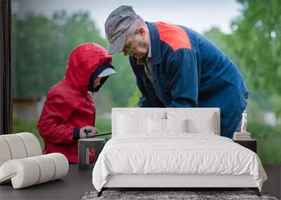 A boy in a red windbreaker and a grandfather in a cap is holding an air gun in summer park. Concept weapons and children, respect, caution, training. Wall mural