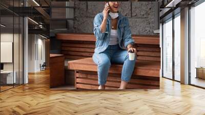 A girl with dark hair in a white medical mask and blue jeans. A young european woman talks on the pnone and drinks coffee on the street. New concept of safety life.  Open wide smile, positive emotions Wall mural