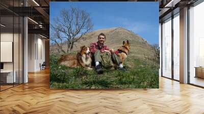 Australian and German Shepherd are traveling with owner in Caucasus. Rest during hike. Young bearded male traveler sitting in clearing among primroses at foot of mountain with two dogs. Wall mural