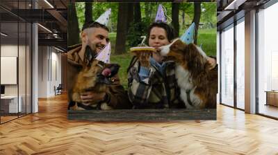 Dogs celebrates birthday with family. Young couple with Australian and German Shepherds in spring green park. Pets birthday party with meat cake and 7 candle, paper hats on heads. Wall mural