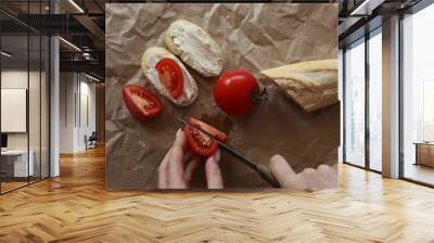 The girl's hands are cutting a tomato on a brown background, next to it lies a sliced baguette, a fresh tomato and two sandwiches. Wall mural