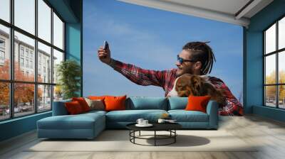 Traveling concept and hiking in mountains with dog. Young Caucasian male blogger in sunglasses sits on top of mountain and takes selfie with Australian Shepherd. Happy pet travel. Wall mural