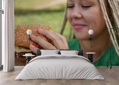 Pretty young woman is eating appetizing hamburger on the street. Close-up Wall mural