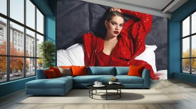 A beautiful slender young luxurious woman in a short red shiny dress lies on white pastel linen in the bed. Advertising fashionable clothes. Advertising of decorative cosmetics.  Wall mural