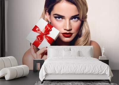 A beautiful young girl with blond hair and red lips is holding a white gift box with a red ribbon. Wall mural