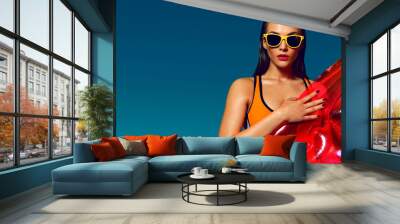 Beautiful tanned girl on the beach against a blue sunny sky. Holds a pink inflatable mattress, sunglasses, wet tanned skin. Rest, south, resort, beach. Sunny day. Wall mural