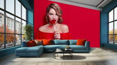 The girl in a red dress on a red background in the studio. Blonde girl holding a red heart-shaped lollipop. Valentine's Day. Advertising. Girl holding a lollipop in his mouth. Wall mural