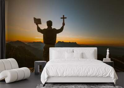 The silhouette of a young man holding a holy bible and raising a christian cross religious symbol in the light and scenery over the sunrise religious background faith concept Wall mural