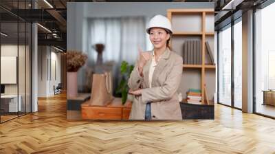 Asian female architect wearing hard hat, standing confidently in office, giving thumbs up. Dressed in beige blazer, emphasizes professionalism, confidence, positive work environment in architecture Wall mural