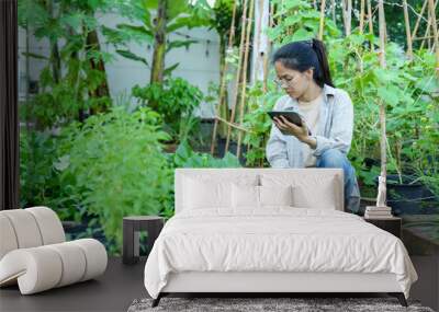 Asian woman using tablet, checking plants in home organic garden with content smile. Surrounded by thriving greenery, embodies commitment to eco-friendly living, gardening, and sustainable practices. Wall mural