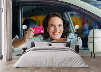 Caucasian woman leaning out of car window smiling and holding pink credit card in hand, parked at an automotive workshop, expressing satisfaction with successful payment for car repair service. Wall mural