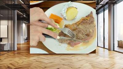 two beautiful hands holding a knife and fork, slicing a large piece of steak in an appetizing dish, mashed potatoes and vegetables on a plate. Wall mural