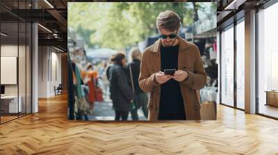 Young man wearing a brown jacket and sunglasses uses his phone at an outdoor market. Wall mural