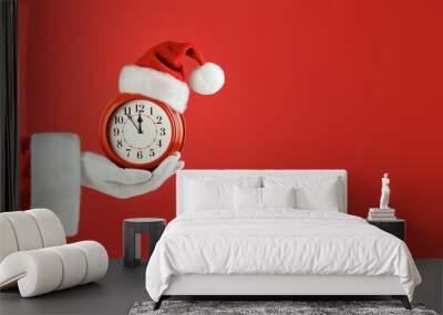 Santa Claus hand holding red alarm clock in Santa hat. Red background. Concept of coming Christmas and New Year, holiday sales. Space for text, banner Wall mural