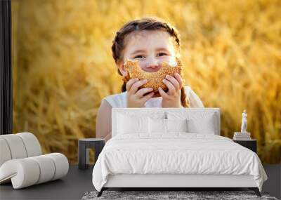 A smiling little girl in a white sundress and two pigtails on a wheat field with a bagel in the open air. He puts a bagel to his mouth in the form of a smile. Wall mural