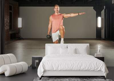 a young Caucasian athlete stands on one leg, moves his arm to the side and raises his leg, and holds a large dumbbell in his other hand. fitness. aerobics. exercises on the mat. physical health Wall mural