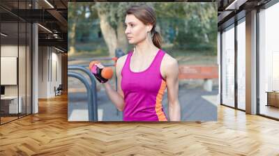 Beautiful slim girl in bright sportswear trains with dumbbell for biceps at outdoor sportground. White earphones, protective gloves. Real woman in morning training, strenght and motivation. Copy space Wall mural