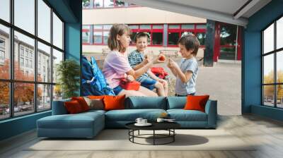 Children sit on a bench in the school yard and eat apples and sandwiches. Snack during break time during class Wall mural