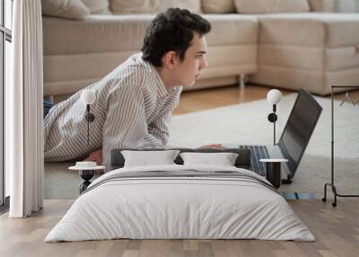 Teenager young man lying on carpet and using laptop at home Wall mural