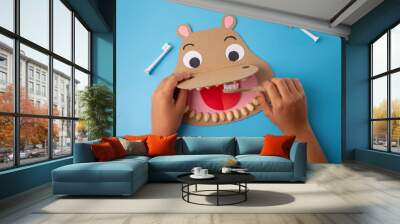 Paper craft hippo head with open mouth, hand holding toothbrush, surrounded by white-handled toothbrushes on blue background, creative and educational dental hygiene project Wall mural