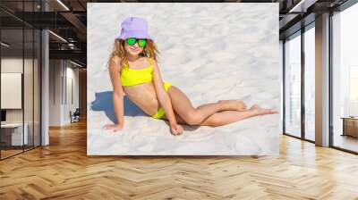 Portrait of a young beautiful model girl with hat and sunglasses posing on the beach. Wearing a bright yellow bikini swimsuit. Summer day, white sand. Wall mural