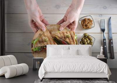 Two halves of a club sandwich on a wooden plate. The plate is held by two female hands. Olives, mustard and cutlery complement the composition. Light wooden background. View from above. Wall mural