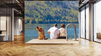 Couple with dog sitting on dock at the lake. Rear view of a couple sitting on pier with dog enjoying stunning view Wall mural