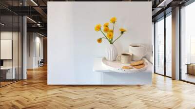 A bouquet of dandelions in a white fluted vase and two cups of different sizes with coffee on a white table. Wall mural