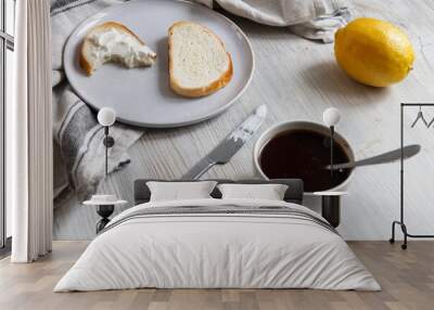 A cup of coffee, two pieces of sugar, a lemon, a piece of bread with curd cheese spread on it on a white ceramic plate with a knife on the table. Wall mural