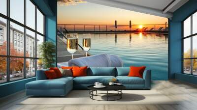 Two glasses of champagne sit beside a rolled towel near a serene pool, as the sun sets, casting vibrant colors over the horizon and creating a peaceful ambiance Wall mural