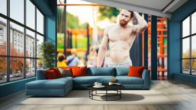 A handsome young Caucasian man with a naked muscular torso with long red hair and a beard posing on a street sports field, an outdoor gym. Sexy guy touches his hair Wall mural