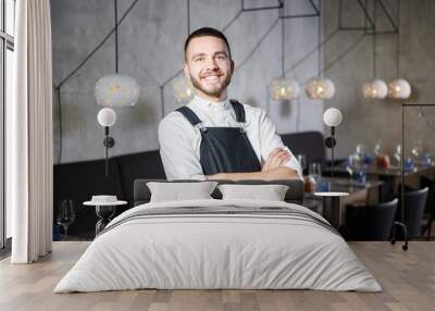 A young, smiling waiter in a restaurant, standing next to the tables with a glass of wine. He wears an apron, looks confidently, folded arms over his chest Wall mural