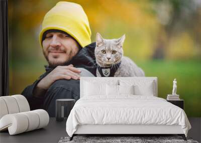 Portrait of tall young Caucasian happy man outdoor playing with gray funny handsome kitten sits on his back on transparent cat backpack carriers bag, close-up nature green background,people and pet Wall mural