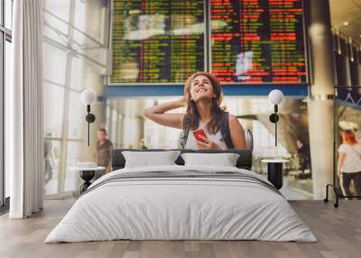 Theme travel and tranosport. Beautiful young caucasian woman in dress and backpack standing inside train station or terminal looking at a schedule holding a red phone, uses communication technology Wall mural