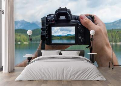 Hands of a male photographer holding a digital camera taking pictures of a idyllic landscape with a lake and mountains while the picture shows at the display Wall mural