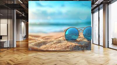 Sunglasses in the sand on a tropical beach with a background of blue ocean and light blue sky. Wall mural