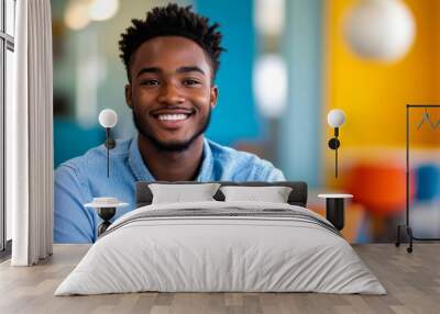 A confident young African American man sits in a lively office environment, smiling broadly while wearing a blue shirt. Bright colors and contemporary design enhance the atmosphere Wall mural