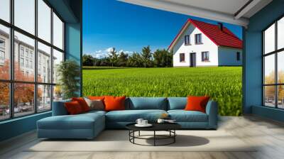 A small white house with a vivid red roof stands proudly on a perfectly manicured green lawn, surrounded by trees under a clear blue sky Wall mural