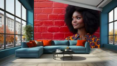 A woman stands against a striking red brick wall, showcasing her voluminous afro and colorful patterned blouse. Her confident pose and the wall's vibrant hue create a bold fashion statement Wall mural